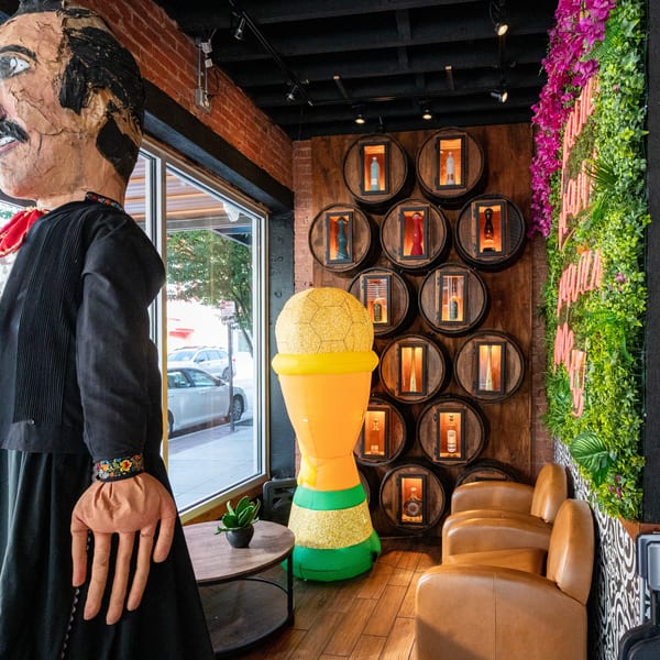 A large paper mache figure stands next to a wall of barrel displays and a green plant wall.