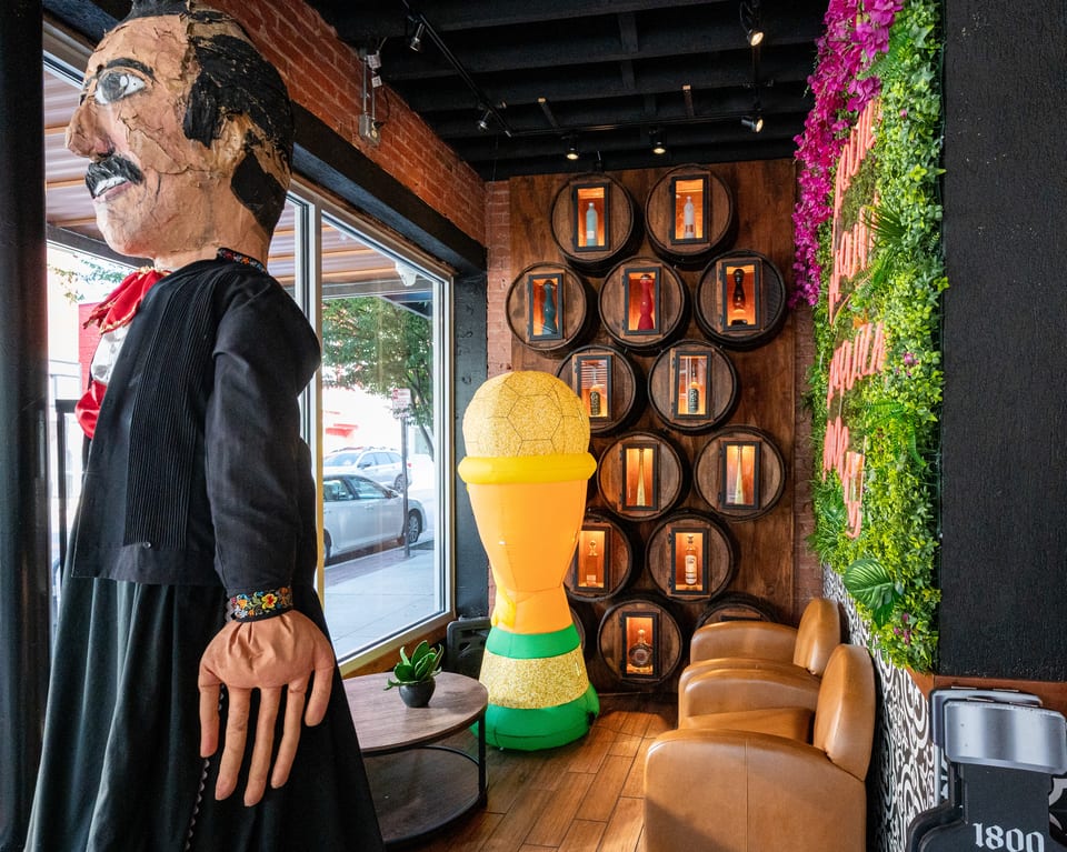 A large paper mache figure stands next to a wall of barrel displays and a green plant wall.