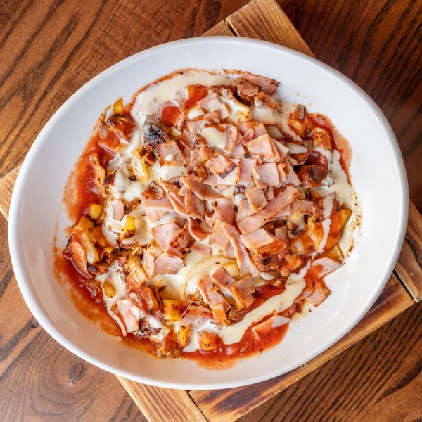 A white oval bowl filled with a dish of ham, melted cheese, and red sauce on a wooden table.
