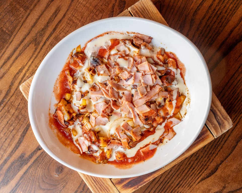 A white oval bowl filled with a dish of ham, melted cheese, and red sauce on a wooden table.