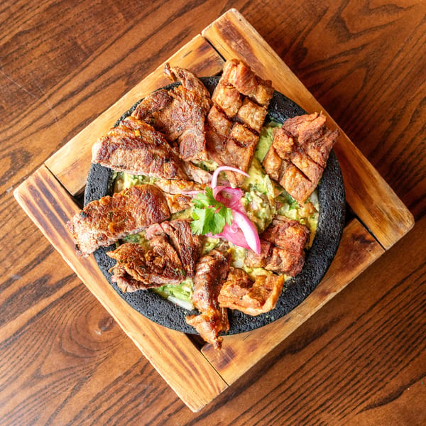 Grilled steak and pork belly over guacamole, garnished with red onion and cilantro, on a wooden board.
