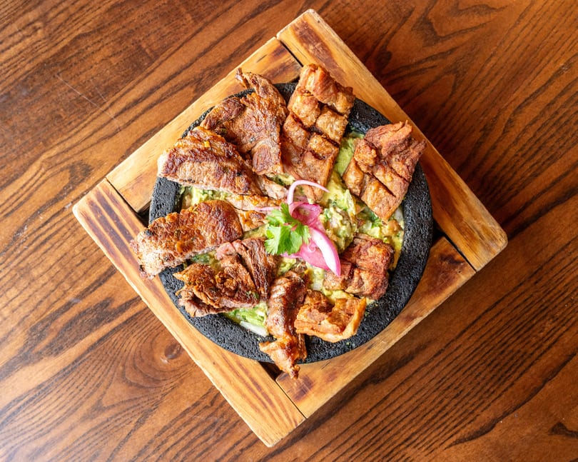 Grilled steak and pork belly over guacamole, garnished with red onion and cilantro, on a wooden board.