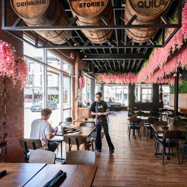 A restaurant interior with a server, a customer, wooden tables, and pink floral decorations hanging from the ceiling.