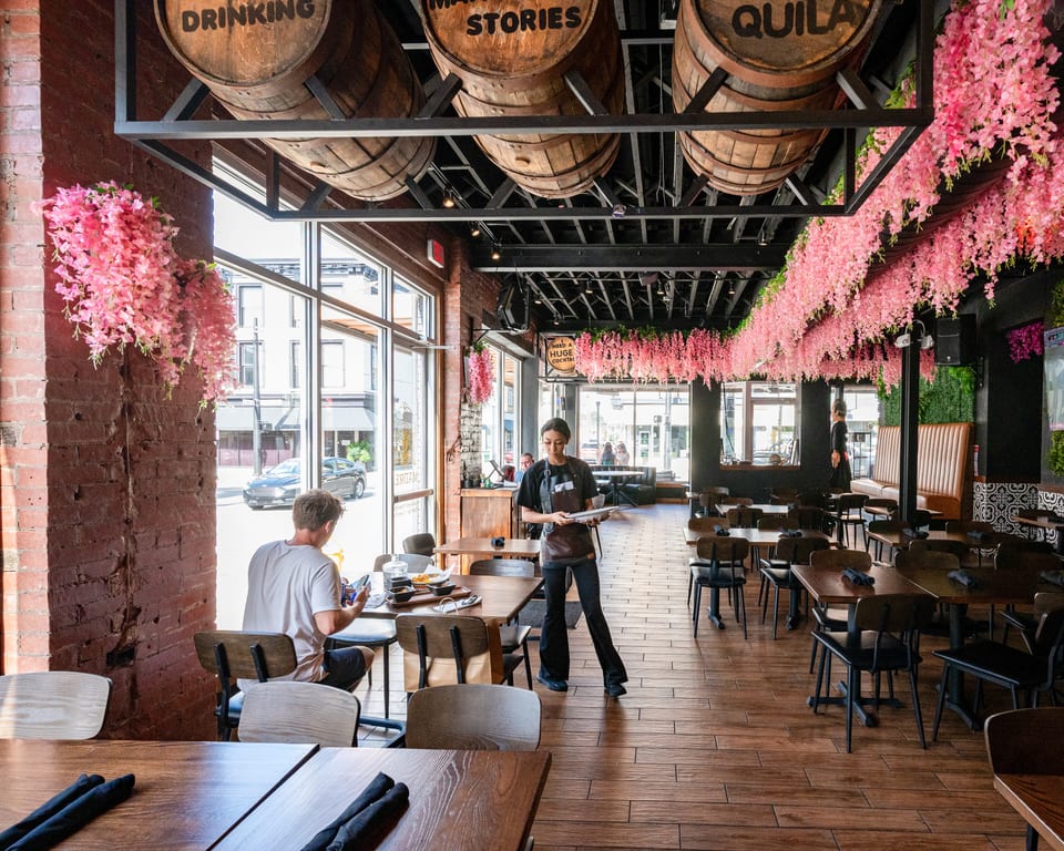 A restaurant interior with a server, a customer, wooden tables, and pink floral decorations hanging from the ceiling.