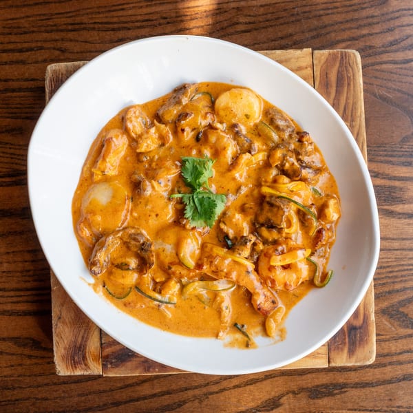 A white bowl of creamy orange curry with shrimp, scallops, and vegetables on a wooden board.