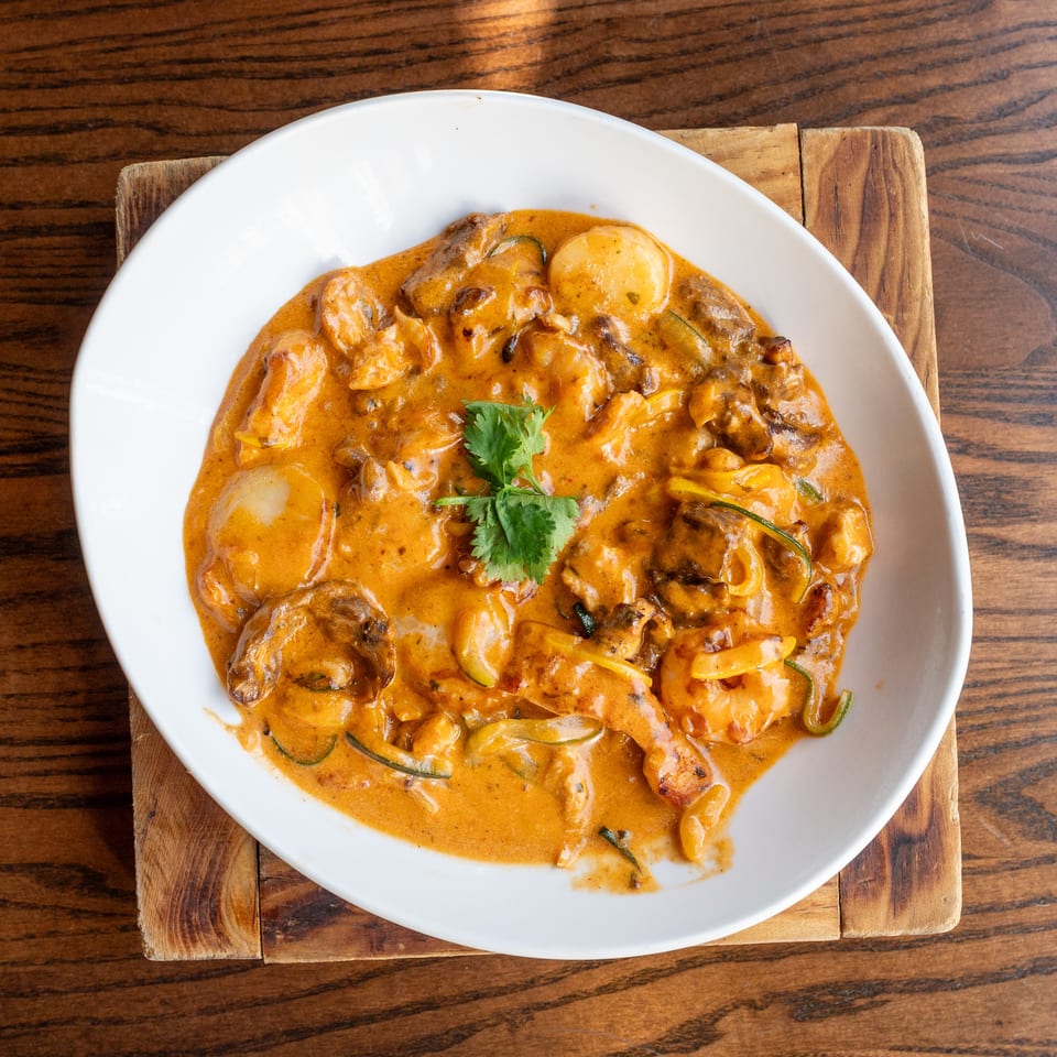 A white bowl of creamy orange curry with shrimp, scallops, and vegetables on a wooden board.