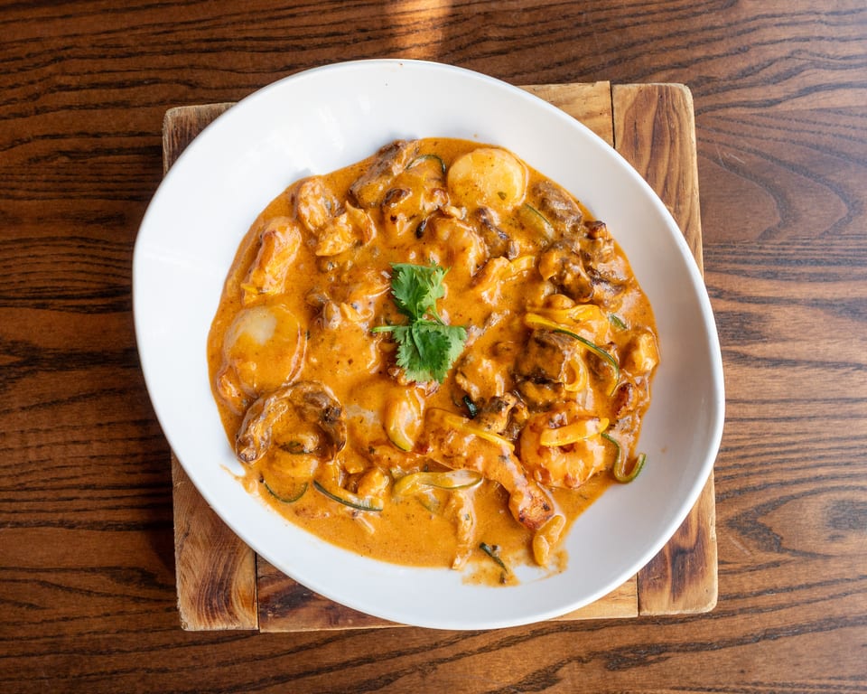 A white bowl of creamy orange curry with shrimp, scallops, and vegetables on a wooden board.