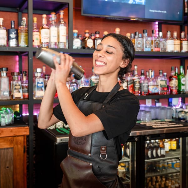 A smiling bartender with closed eyes shakes a cocktail shaker behind a well-stocked bar.