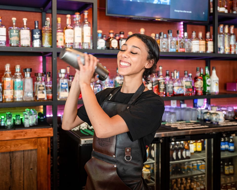 A smiling bartender with closed eyes shakes a cocktail shaker behind a well-stocked bar.