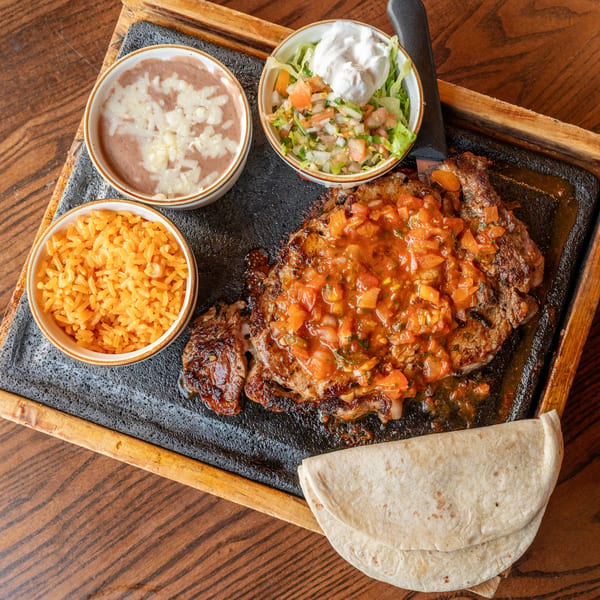 Grilled steak with tomato salsa, rice, refried beans, salad, and tortillas on a wooden tray.