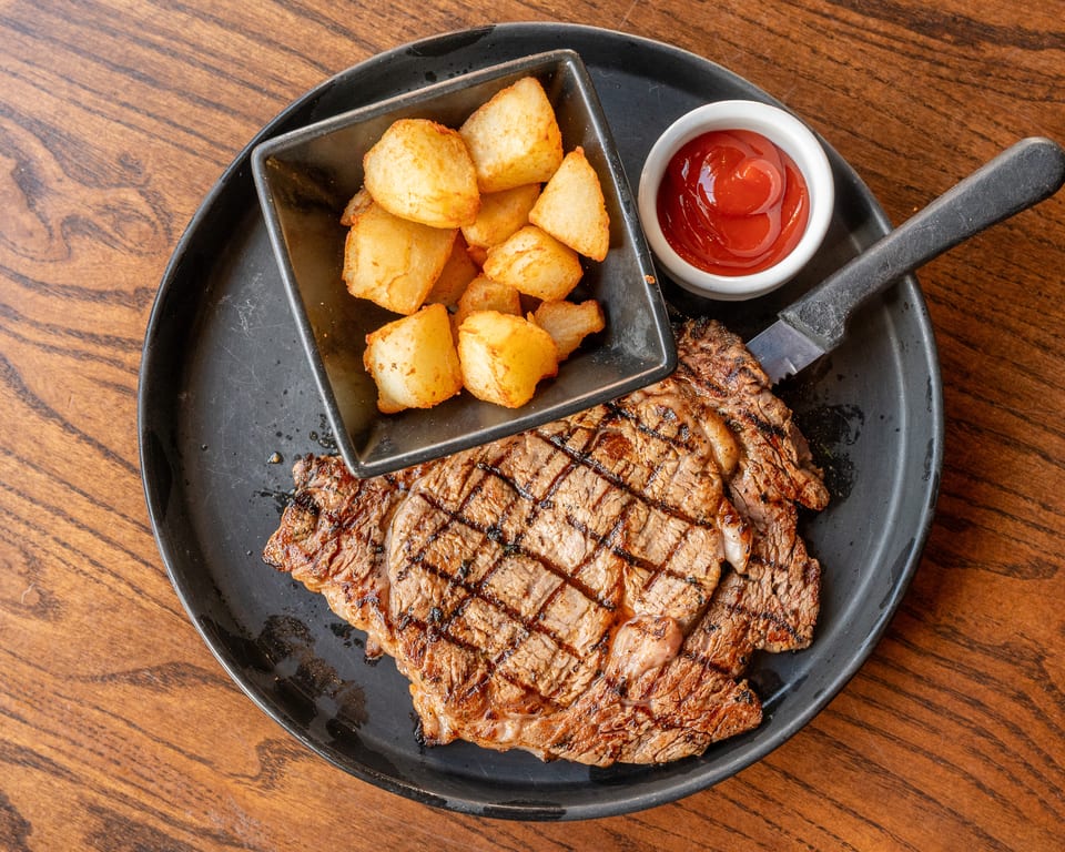 Grilled steak with hash browns and a side of ketchup on a black plate.