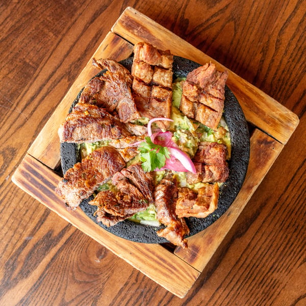 Grilled meat and pork belly served on a bed of guacamole in a black stone bowl.