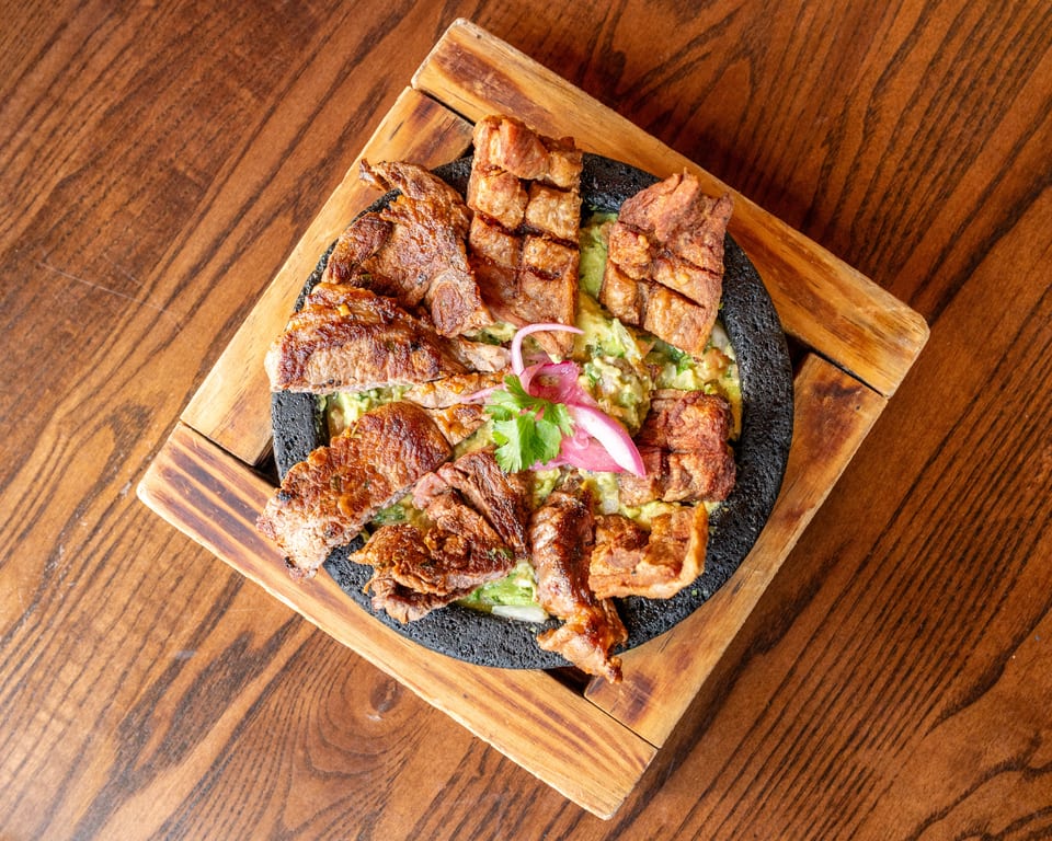 Grilled meat and pork belly served on a bed of guacamole in a black stone bowl.