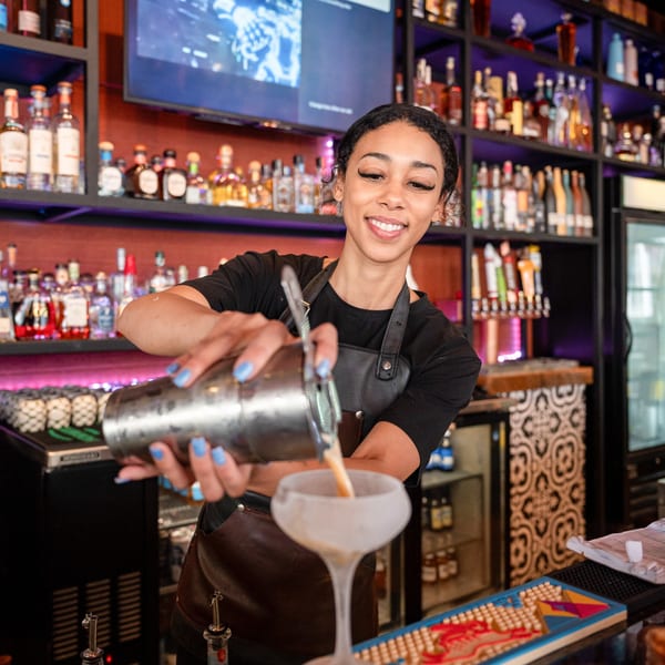 Smiling bartender pouring a cocktail from a shaker into a frosted glass at a bar.