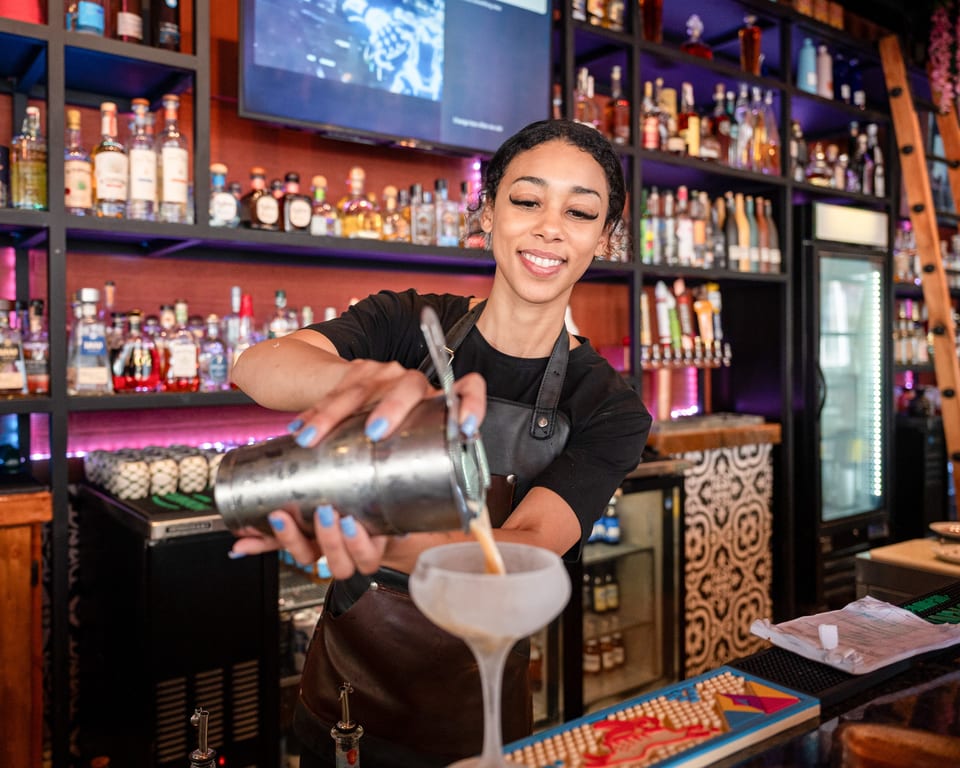 Smiling bartender pouring a cocktail from a shaker into a frosted glass at a bar.