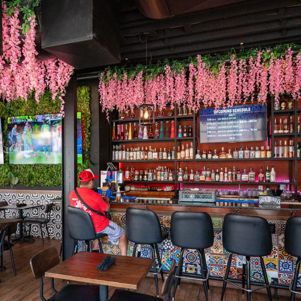 A vibrant bar with pink floral decor, a man at the counter, and TVs displaying a soccer match.