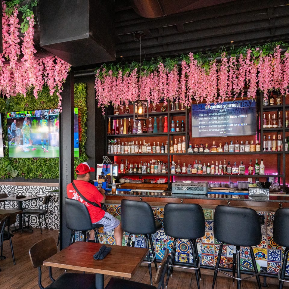 A vibrant bar with pink floral decor, a man at the counter, and TVs displaying a soccer match.