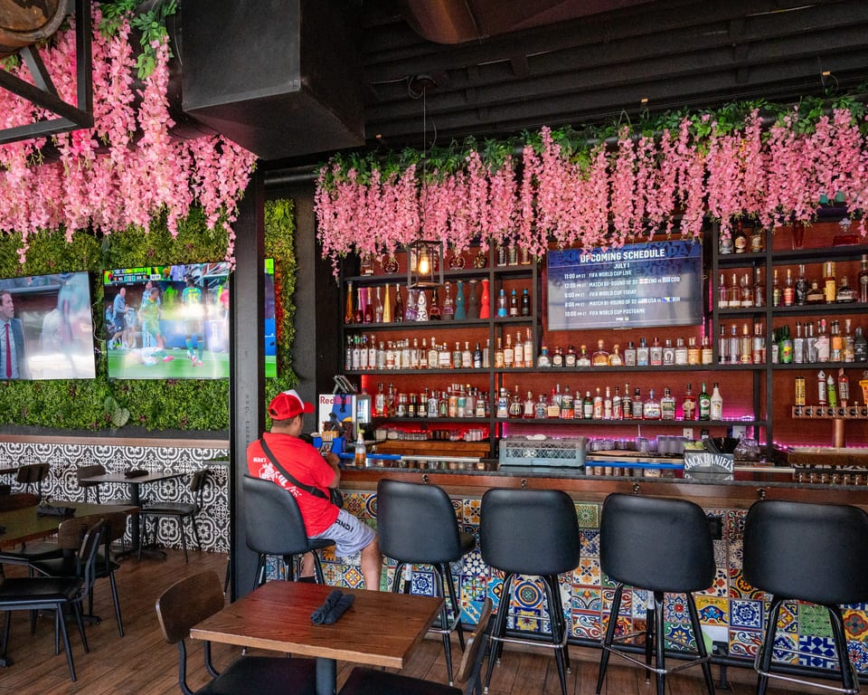 A vibrant bar with pink floral decor, a man at the counter, and TVs displaying a soccer match.