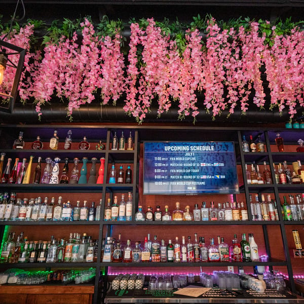 A bar with shelves of liquor bottles, a TV displaying a FIFA World Cup schedule, and pink floral decor.