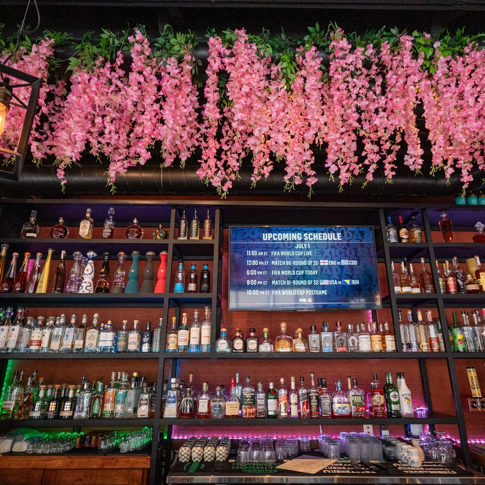 A bar with shelves of liquor bottles, a TV displaying a FIFA World Cup schedule, and pink floral decor.