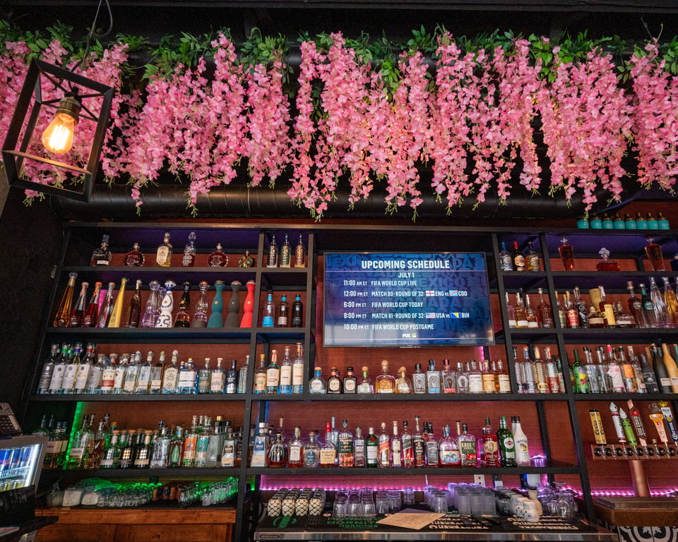 A bar with shelves of liquor bottles, a TV displaying a FIFA World Cup schedule, and pink floral decor.