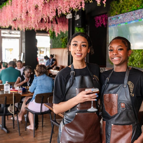 Two smiling female servers in black and brown aprons, one holding a cocktail, in a vibrant restaurant.