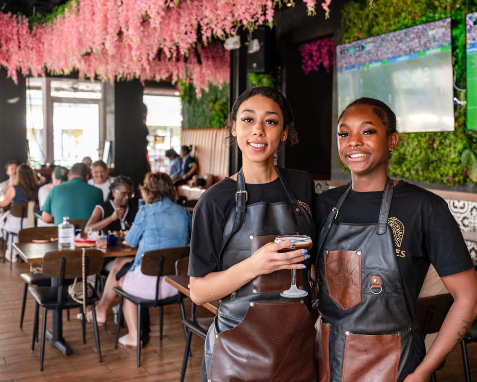 Two smiling female servers in black and brown aprons, one holding a cocktail, in a vibrant restaurant.