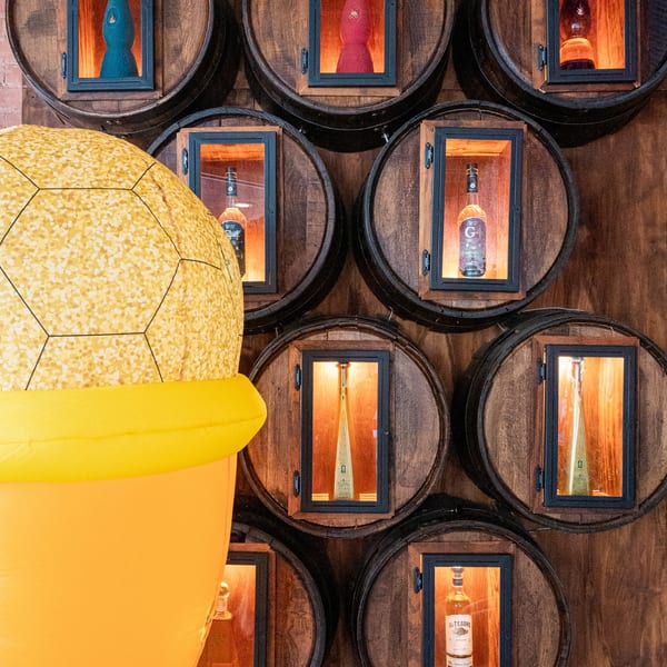 A large inflatable ice cream cone stands beside a wall of wooden barrels displaying liquor bottles.