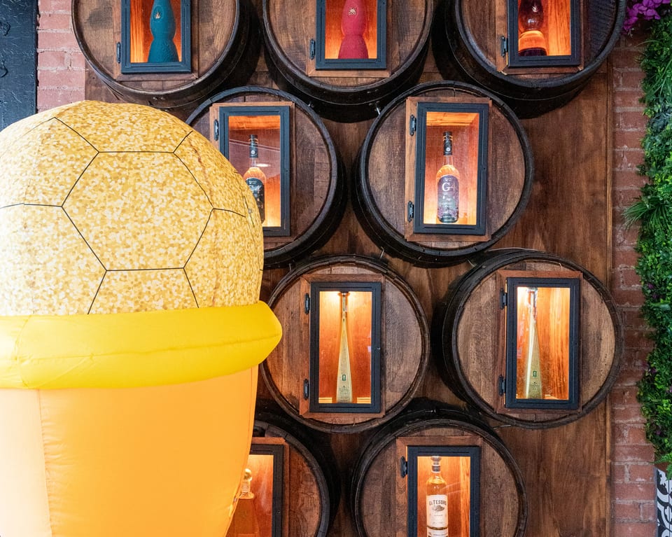 A large inflatable ice cream cone stands beside a wall of wooden barrels displaying liquor bottles.