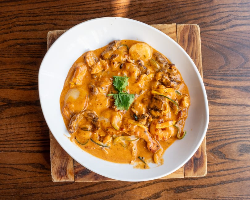 A white bowl of creamy orange curry with shrimp, scallops, and vegetables on a wooden table.