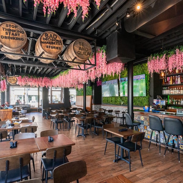 Vibrant restaurant interior with pink floral decor, wooden tables, a bar, and barrels with playful sayings.