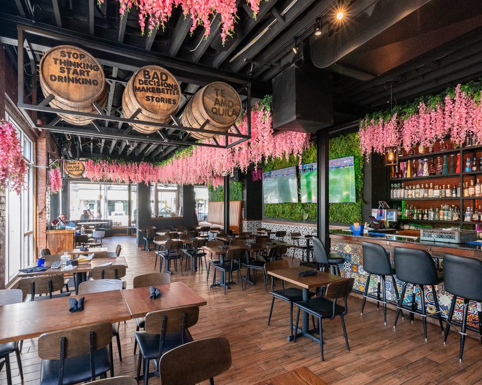 Vibrant restaurant interior with pink floral decor, wooden tables, a bar, and barrels with playful sayings.