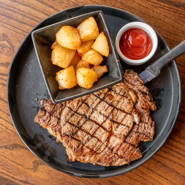 Grilled steak with crisscross sear marks, a side of roasted potatoes, and ketchup on a black plate.