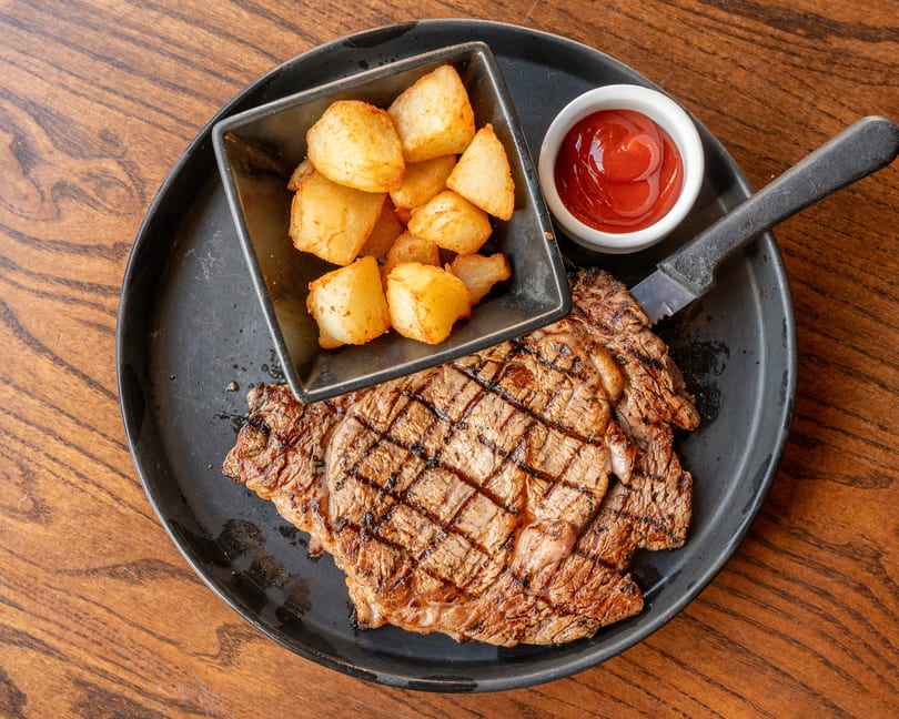 Grilled steak with crisscross sear marks, a side of roasted potatoes, and ketchup on a black plate.