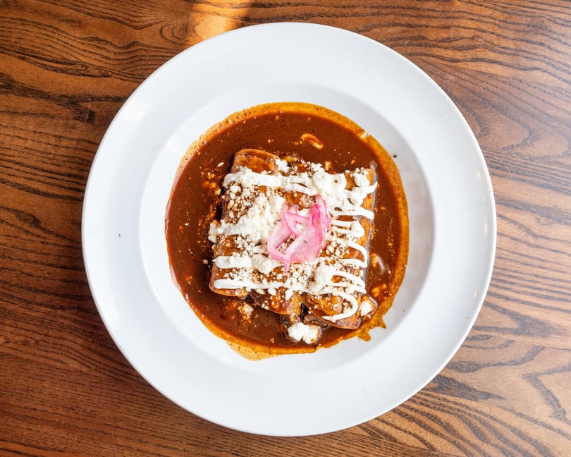 Enchiladas covered in rich mole sauce, crumbled cheese, crema, and pickled red onions on a white plate.
