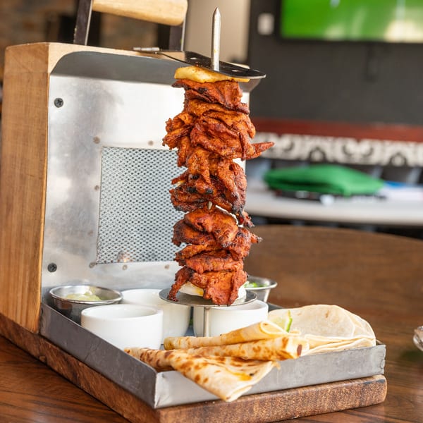 Al pastor meat on a vertical spit with tortillas and sauces on a wooden tray.