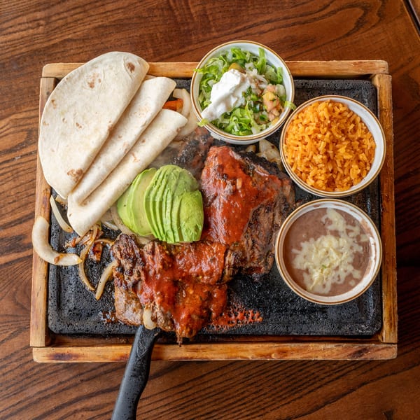 Sizzling fajita platter with steak, tortillas, avocado, rice, beans, and salad on a wooden table.