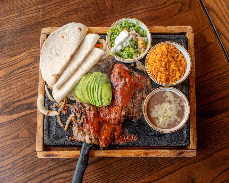 Sizzling fajita platter with steak, tortillas, avocado, rice, beans, and salad on a wooden table.
